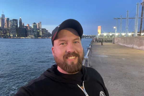 photo of Thomas Lupton wearing a hat, beard, and standing on a pier in Brooklyn with the East River and NYC skyline in the background